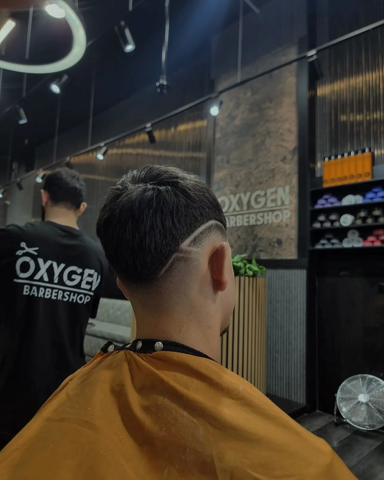 Hard-part fade in progress — a barber in Oxygen tee working behind a seated client in the brass chair